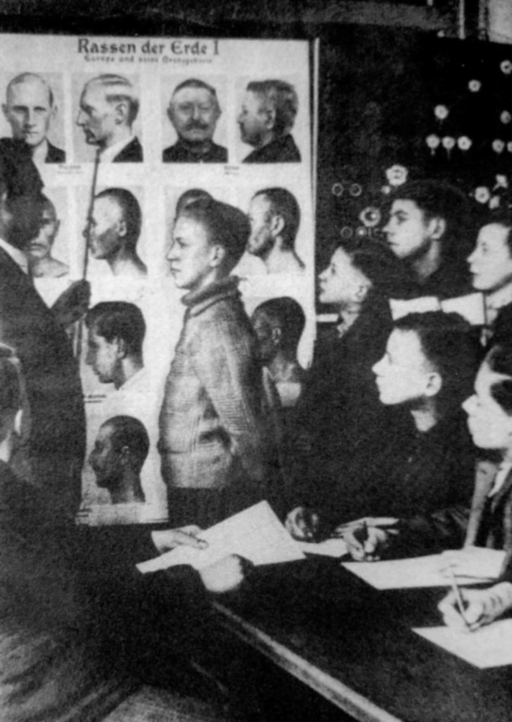 Photograph showing a boy standing beside a poster with multiple faces at various angles. A man stands in front of the boy and points at the poster with a rod while five other children observe.